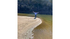 A person standing on a sandy beach with arms outstretched, facing a body of water and a forested hill in the background. - Brazos River Ranch near PK & Rocker B Ranch  (Mineral Wells)