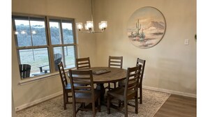A dining area with a round table and wooden chairs, a chandelier, a large window with a view, and a desert-themed painting on the wall. - Brazos River Ranch near PK & Rocker B Ranch  (Mineral Wells)