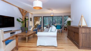 A modern living room with a wooden dining table, a white sofa, and a TV mounted on the wall. - Wooden House With Heated Pool, Fiber Internet and View Into Forest (Moliets-et-Maa)