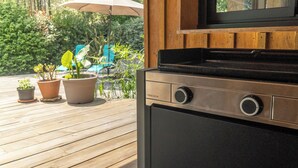 A kitchen area with a built-in stove, wooden deck, potted plants, and a patio umbrella. - Wooden House With Heated Pool, Fiber Internet and View Into Forest (Moliets-et-Maa)