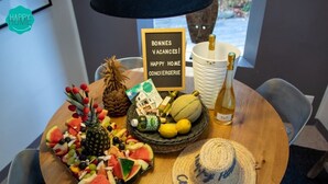 Una mesa con una bandeja de frutas, una botella de vino, una cesta de pan, un sombrero y un cartel con un mensaje.