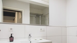 Ein Badezimmer mit einem weißen Waschbecken, einem Spiegel und einer in der Spiegelung sichtbaren Dusche.
