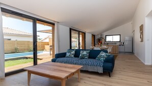 Ein modernes Wohnzimmer mit einem Holztisch, einem blauen Sofa mit gemusterten Kissen und Blick auf einen Garten mit Pool.