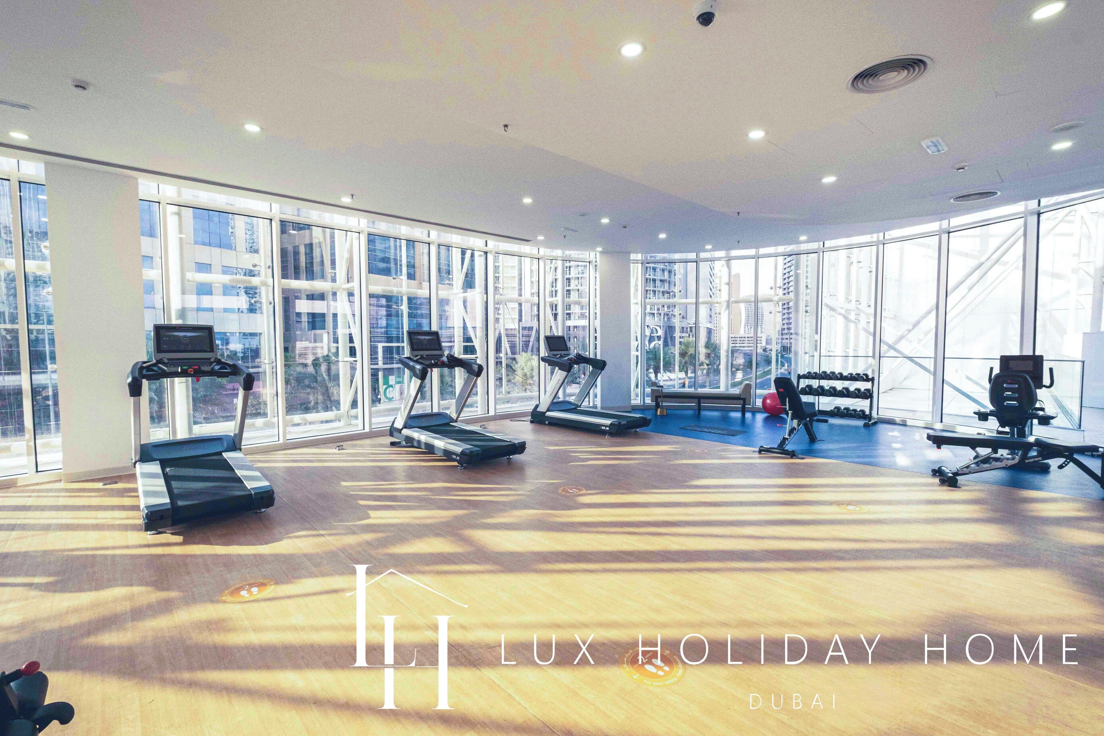 A spacious gym with treadmills, a rowing machine, and a weight bench. Large windows offer views of the city.