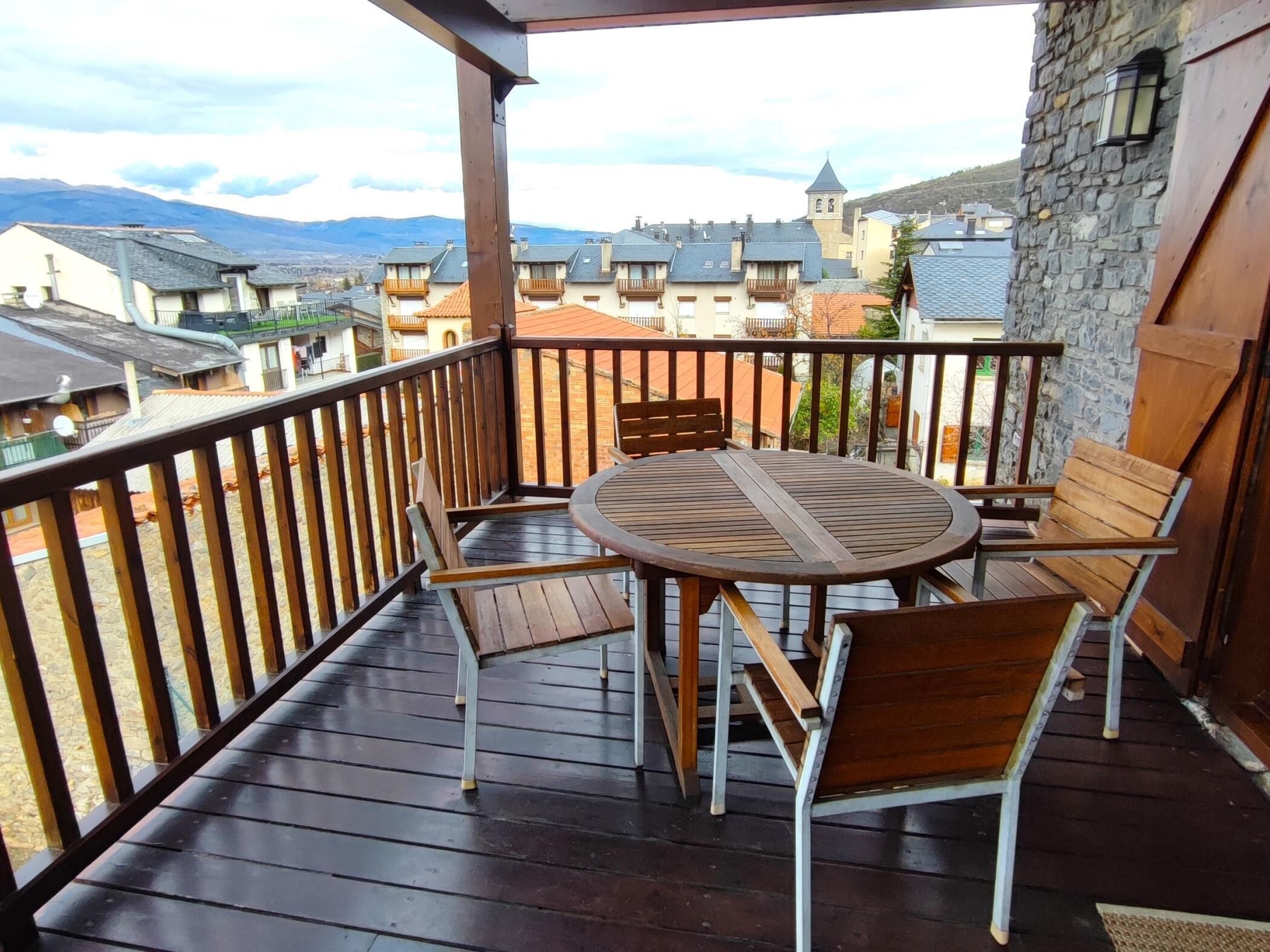 A wooden deck with a round table and chairs, overlooking a town with stone buildings and a mountain in the background.