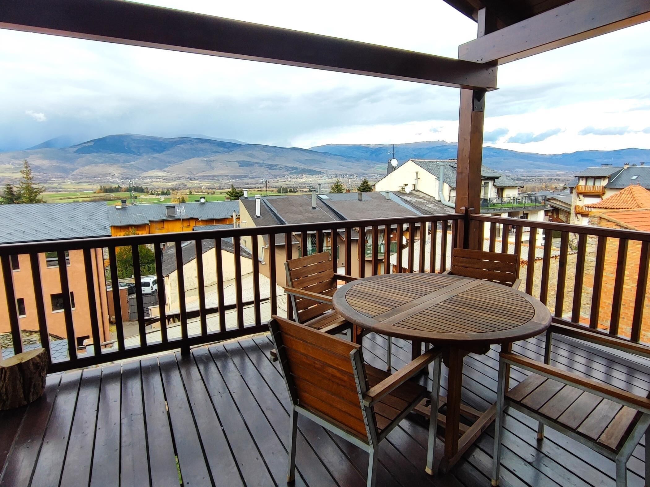 A wooden deck with a round table and chairs, overlooking a residential area with houses and mountains in the distance.