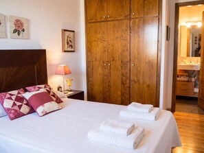 A hotel room with a bed, pillows, a nightstand, a lamp, a wooden wardrobe, and a bathroom visible through an open door.