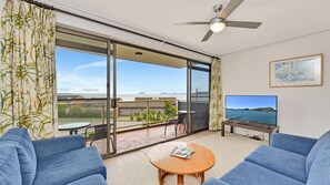A spacious living room with a large sliding glass door leading to a balcony, a ceiling fan, a flat-screen TV, and a framed picture on the wall. - Triview - Mitchell Street, Unit 4/1-3, Soldiers Point - Spectacular water views (Soldiers Point)