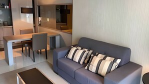 A modern living room with a grey sofa, striped cushions, a dining table with chairs, and a kitchen area in the background. - Brisas do Lago - apartamento 7 (Brasília)