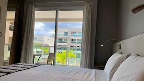 A hotel room with a large bed, a view of buildings and trees through the window, and a balcony with a chair. - Brisas do Lago - apartamento 7 (Brasília)