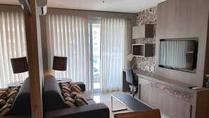 A modern hotel room with a sofa, a small table, a TV, and a balcony with curtains. - Brisas do Lago - apartamento 7 (Brasília)