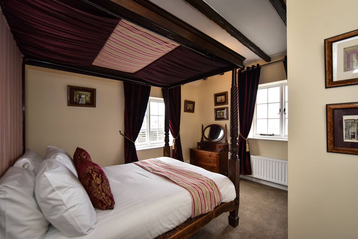 Een slaapkamer met een hemelbed, een houten dressoir en ingelijste foto’s aan de muur.