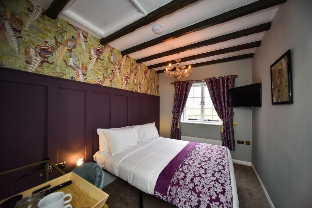 A bedroom with a bed, a wooden side table with cups and a remote, a window with curtains, and a TV on the wall.