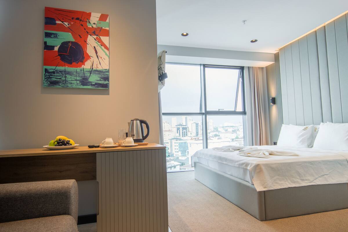 A modern hotel room with a large bed, a desk with a kettle, a fruit bowl, and a city view through the window.