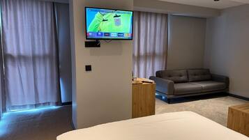 Quarto de hotel moderno com uma cama grande, um sofá, uma TV fixada na parede e uma janela com cortinas.