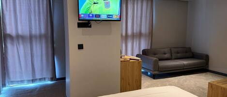 Quarto de hotel moderno com uma cama grande, um sofá, uma TV fixada na parede e uma janela com cortinas.