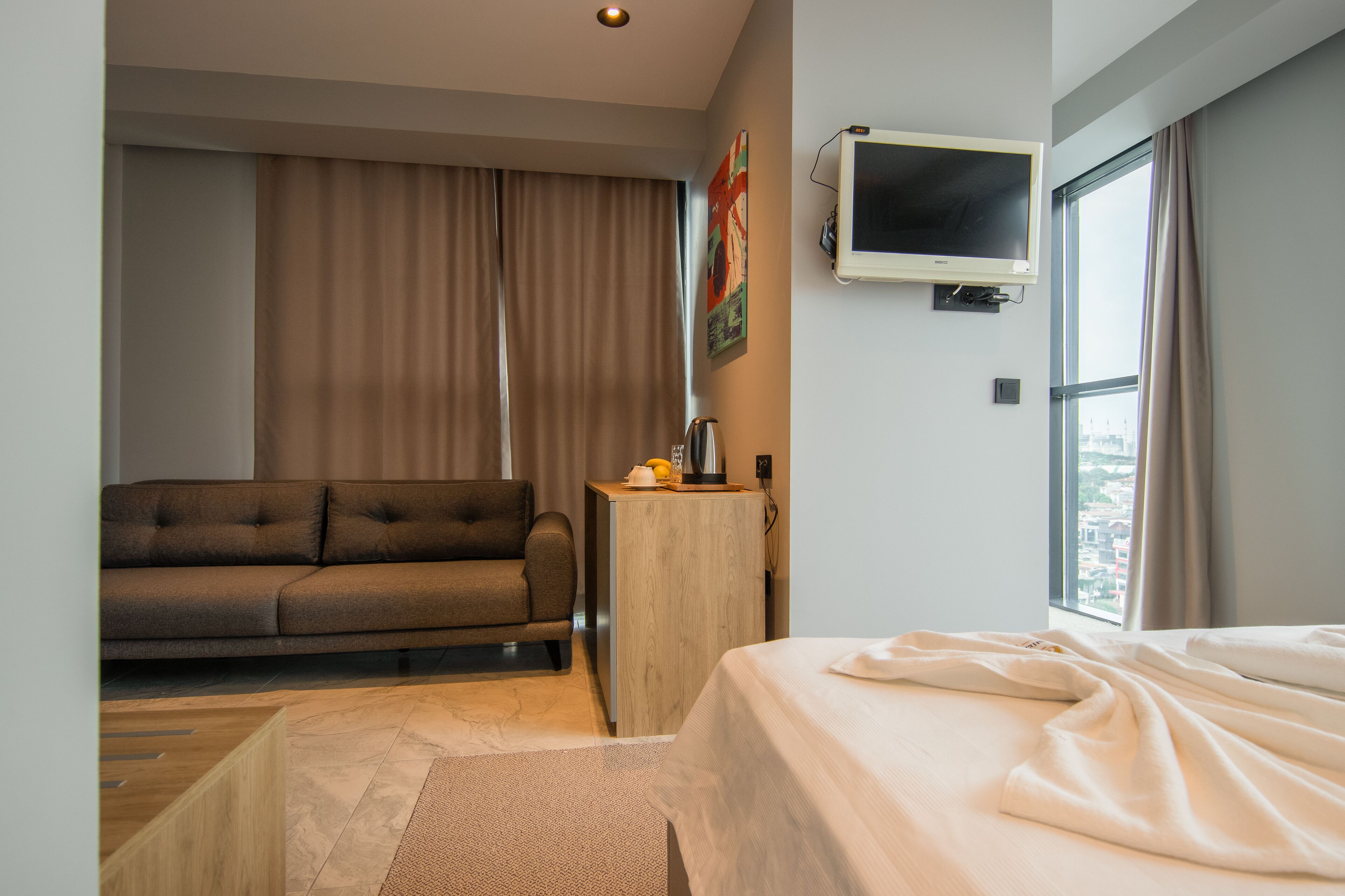 Una moderna camera d'hotel con un letto, un divano, un tavolino con una lampada, una TV a parete e una finestra con tende.
