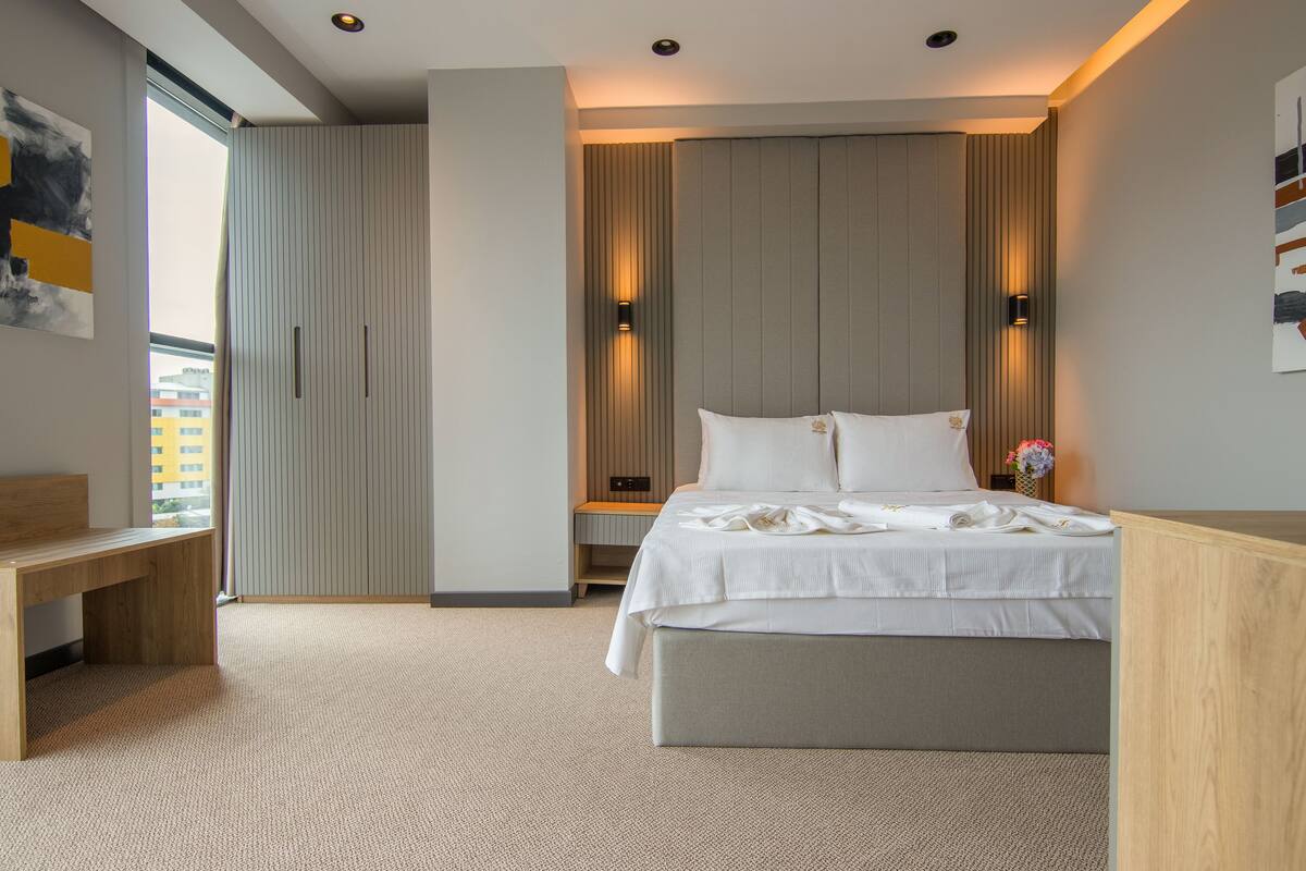 A modern bedroom with a large bed, a wooden desk, and a view of the city through a window.