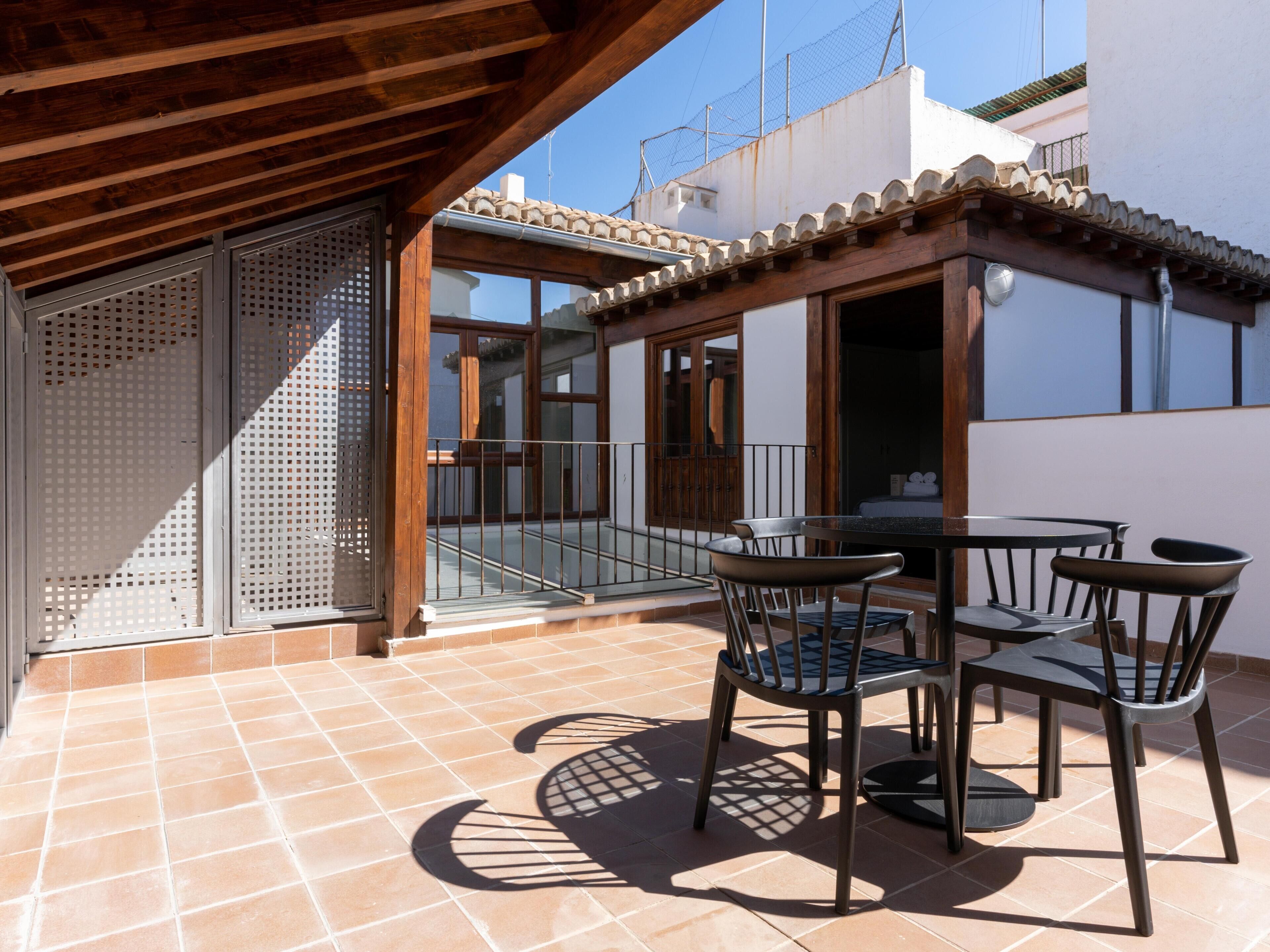 Un patio exterior con una pérgola de madera, un ventanal de vidrio que da a un balcón y una mesa con sillas.