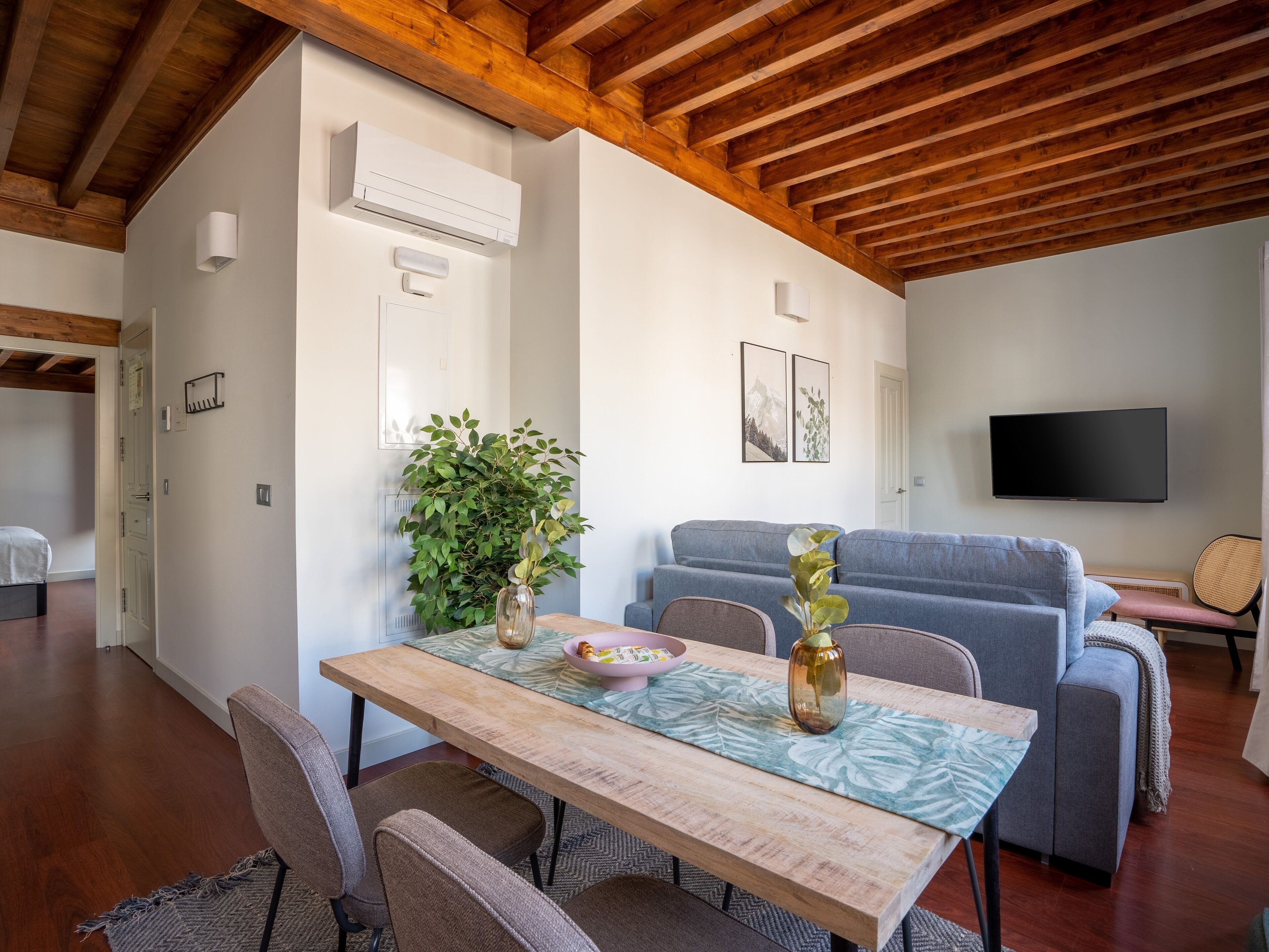 A modern living room with a wooden ceiling, a grey sofa, a TV mounted on the wall, and a dining area with a wooden table and chairs.