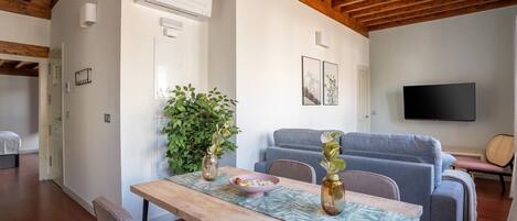A modern living room with a wooden ceiling, a grey sofa, a TV mounted on the wall, and a dining area with a wooden table and chairs.