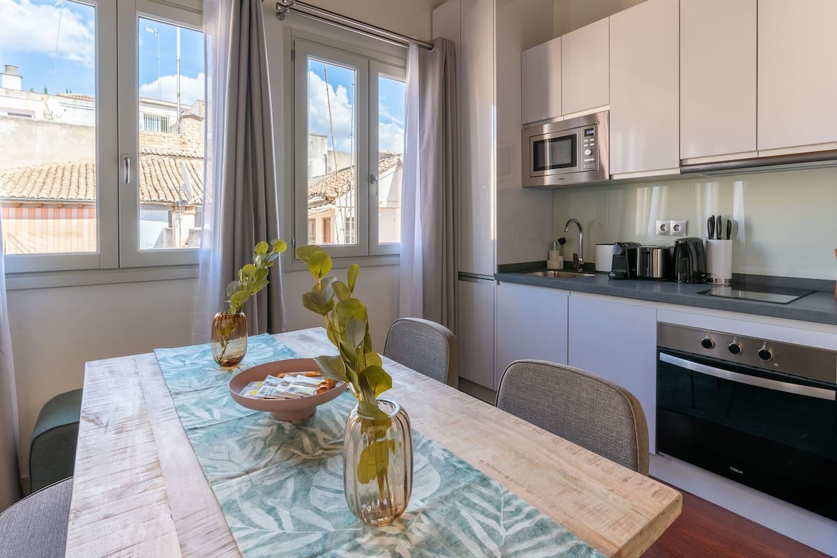 Una cocina moderna con armarios blancos, un horno de acero inoxidable y un microondas. Hay una mesa de comedor con un jarrón y un tazón.