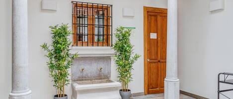 A courtyard with a fountain, two potted plants, a wooden door, and a window with bars.