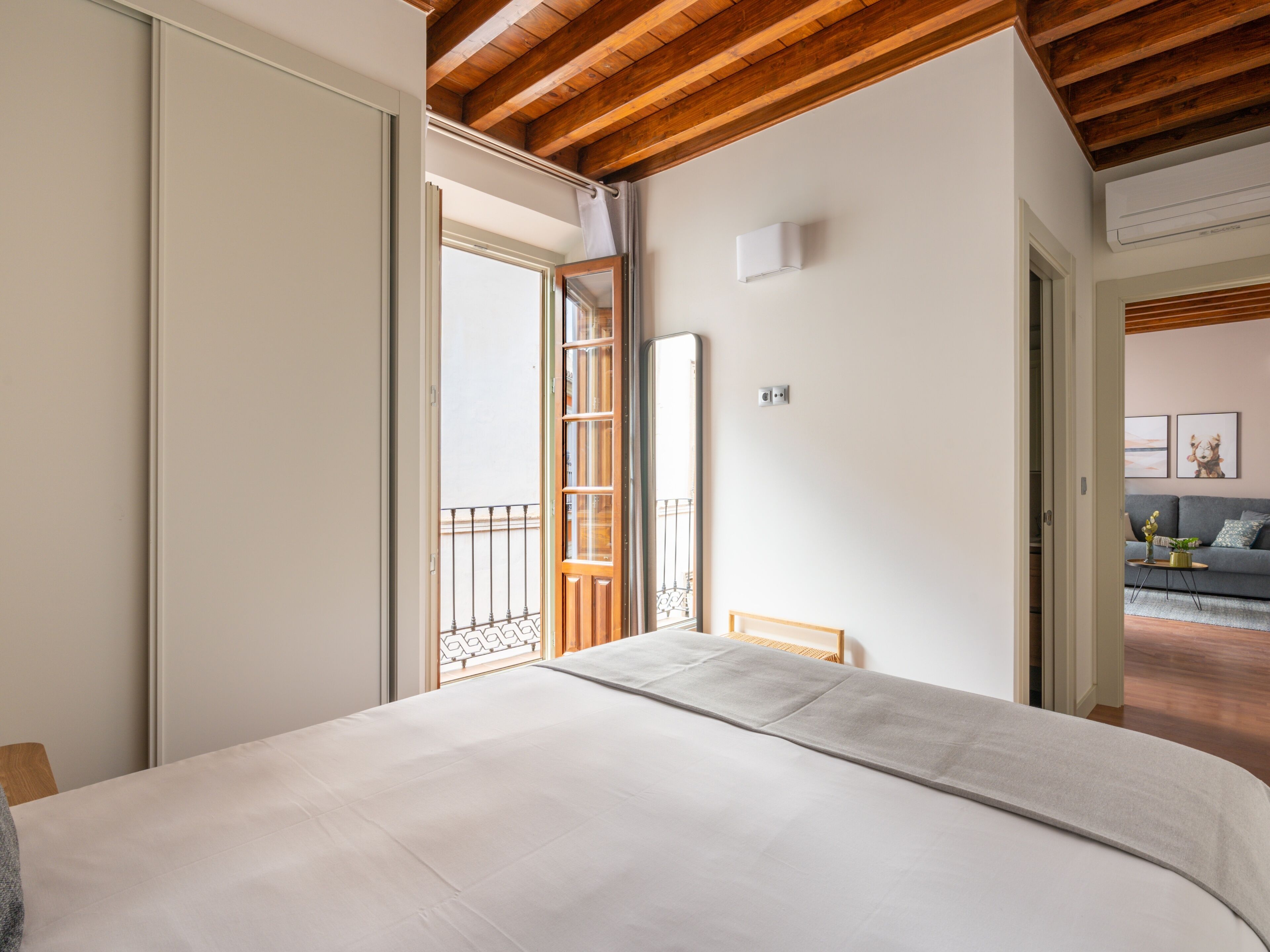 Une chambre dotée d’un grand lit, de poutres apparentes en bois au plafond, d’une porte coulissante donnant sur un balcon et d’un canapé dans le salon attenant.