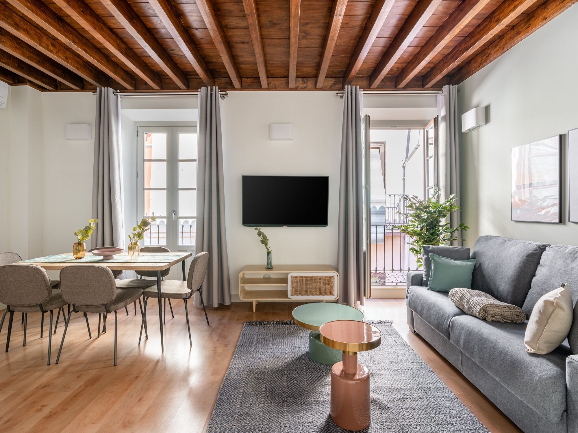Ein modernes Wohnzimmer mit Holzboden, einem grauen Sofa, einem Essbereich mit Tisch und Stühlen sowie einem an der Wand befestigten Fernseher.