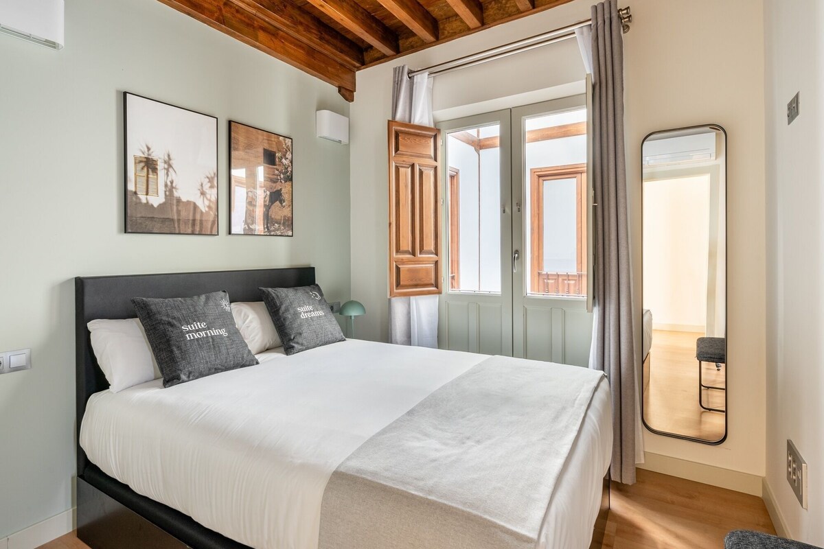 Een slaapkamer met een bed, houten plafond, een raam met gordijnen, een spiegel en ingelijste foto's aan de muur.