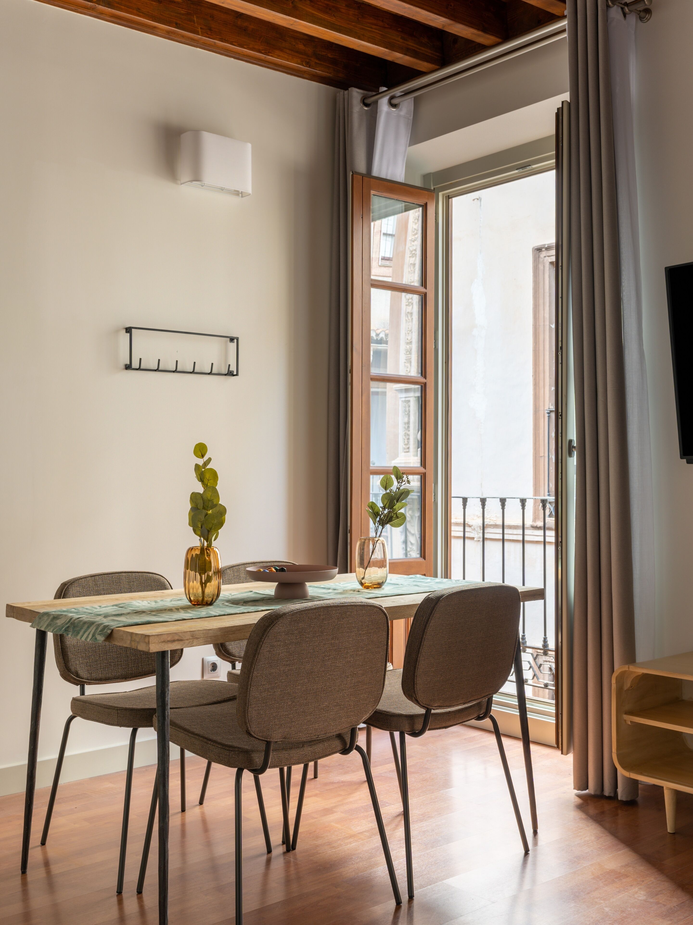 Comedor con mesa de madera, cuatro sillas, un florero con hojas verdes y una ventana con cortinas.