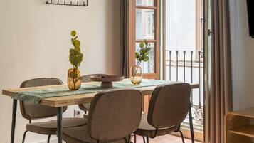 Comedor con mesa de madera, cuatro sillas, un florero con hojas verdes y una ventana con cortinas.
