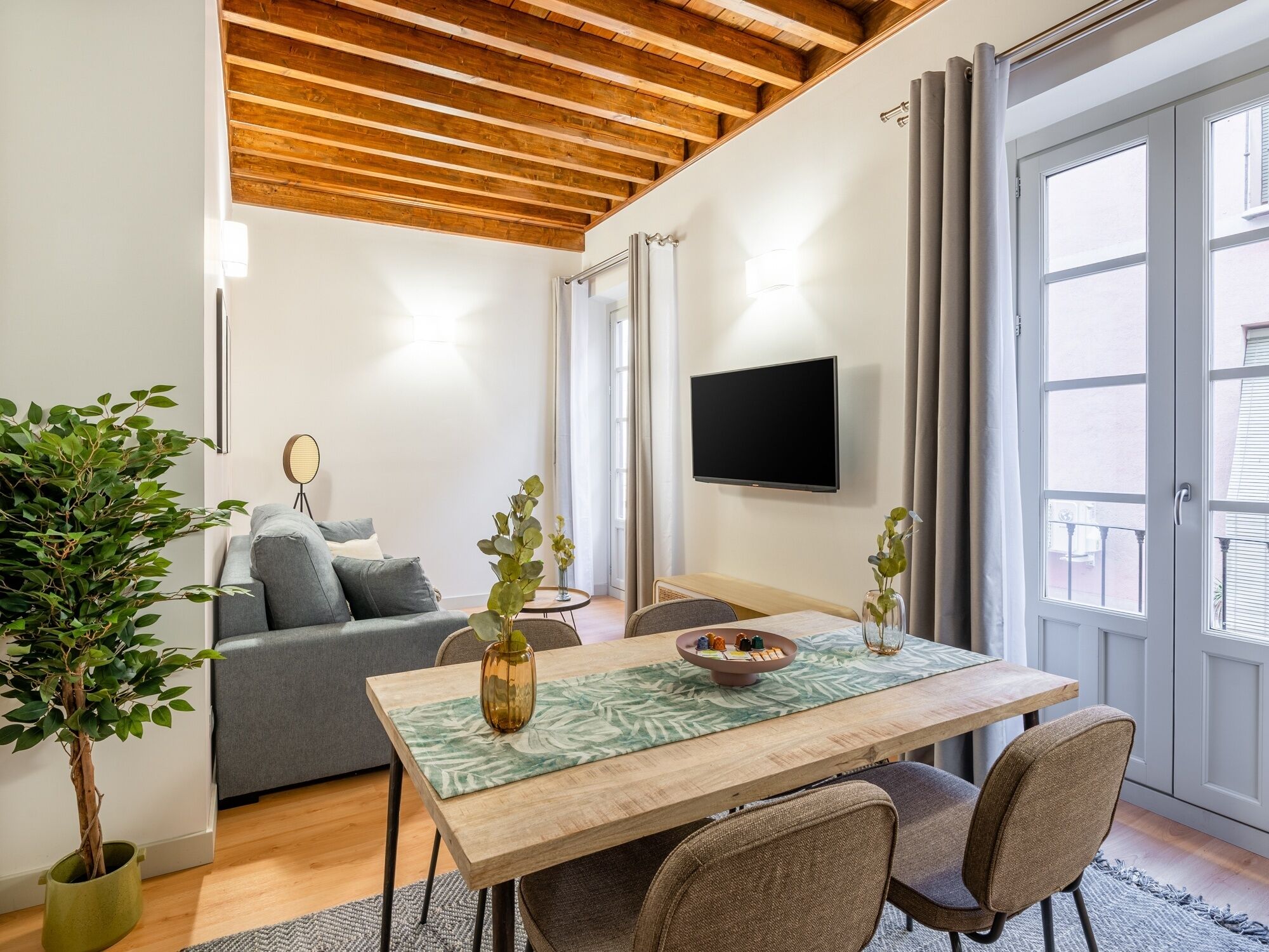 Una sala de estar moderna con techo de madera, un sofá gris, una mesa de comedor con sillas, un televisor montado en la pared y un ventanal con cortinas.