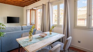 Una sala de estar moderna con techo de madera, un sofá gris, una mesa de comedor con sillas, un televisor montado en la pared y ventanas grandes con cortinas.