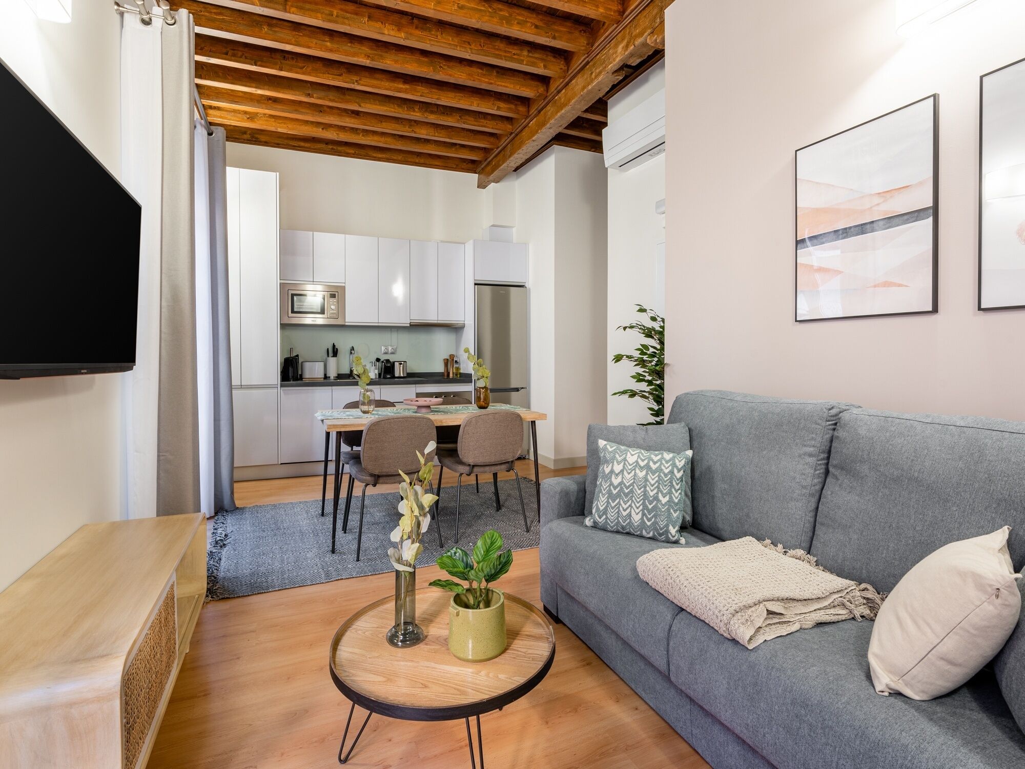 A modern living room with a grey sofa, a coffee table with a vase and plant, a flat-screen TV, and a wooden ceiling.