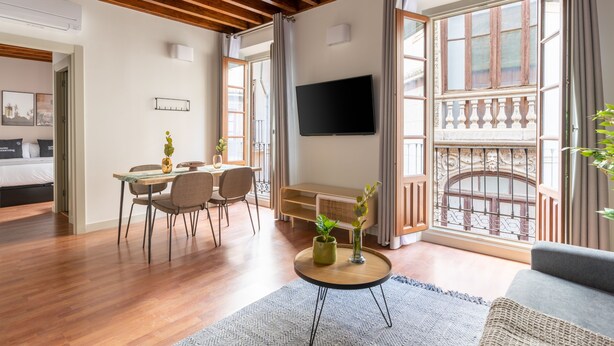 Una sala de estar moderna con piso de madera, un área de comedor con sillas, una mesa de centro con plantas, un televisor montado en la pared y ventanas amplias con vista al balcón.