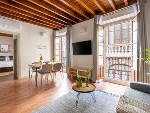 Un salon moderne avec un plancher en bois, un coin repas avec des chaises, une table basse agrémentée de plantes, un téléviseur fixé au mur et de grandes fenêtres donnant sur un balcon.