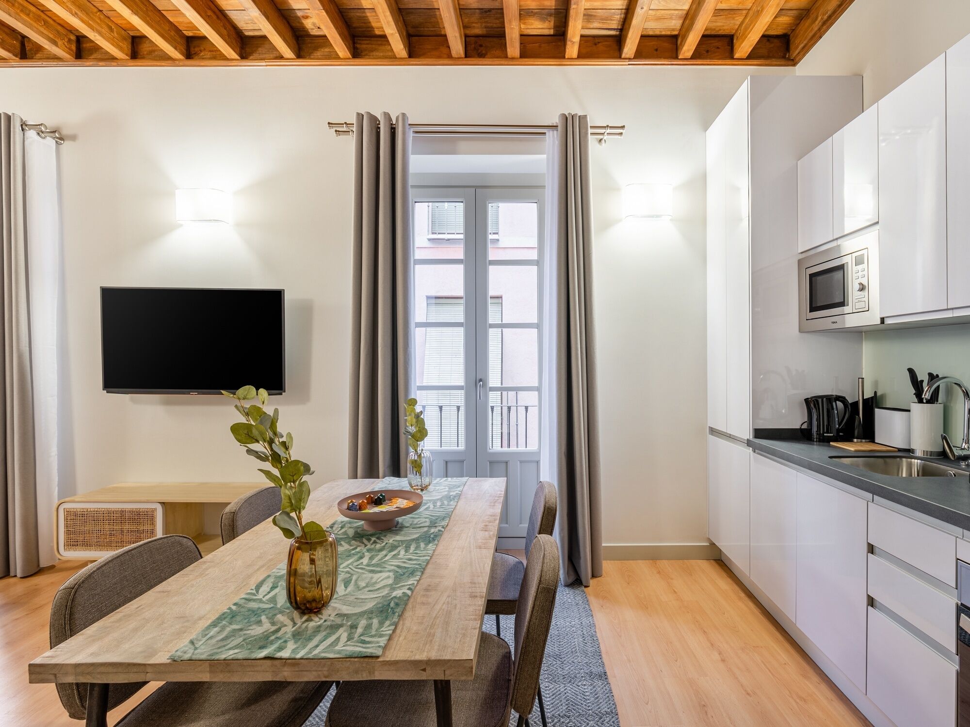 A modern kitchen with white cabinets, a microwave, and a sink. There is a dining table with chairs and a vase of flowers. A television is mounted on the wall.