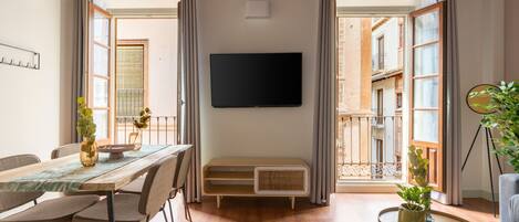 A modern living room with a wooden ceiling, a flat-screen TV mounted on the wall, a dining table with chairs, a small side table with a plant, and a view of the outside balcony.