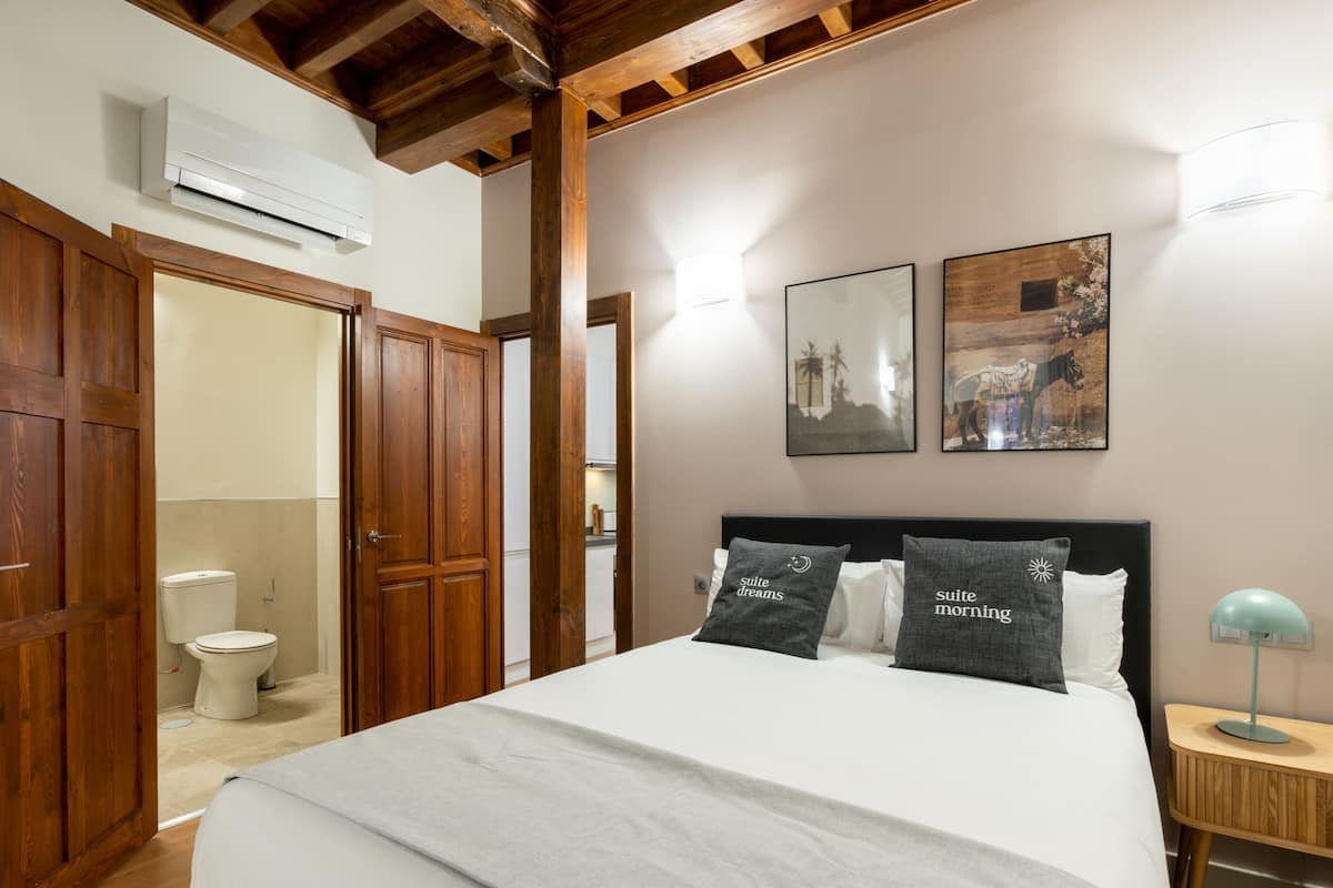 Un dormitorio con cama, mesita de noche, lámpara, armario de madera y baño visible a través de una puerta abierta.