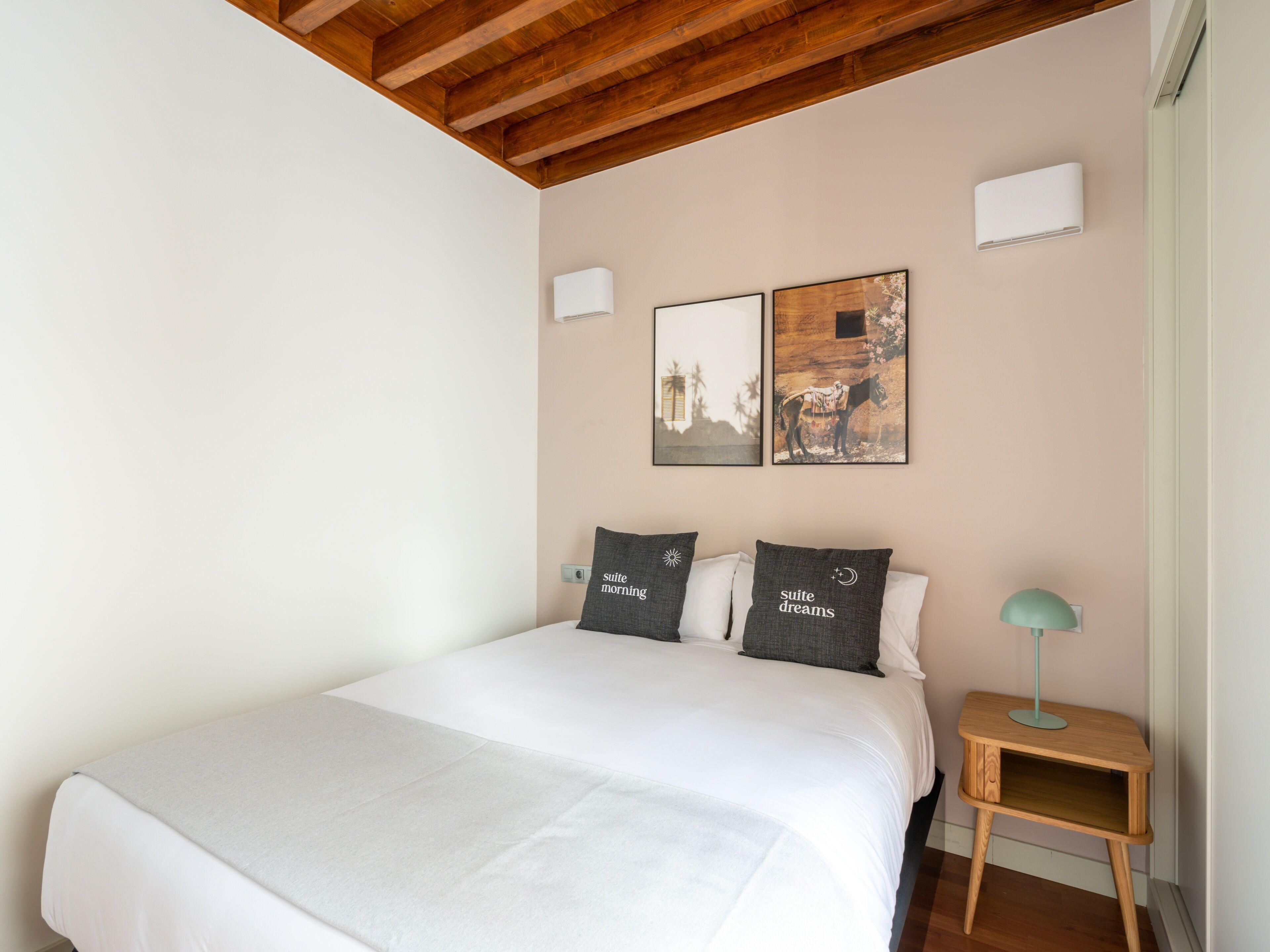 A bedroom with a wooden ceiling, a bed with white bedding, two pillows with text, a small bedside table with a lamp, and two framed pictures on the wall.