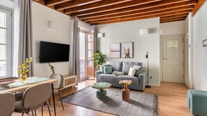 Una sala de estar moderna con techo de madera, un sofá gris, una mesa de comedor con sillas, un televisor montado en la pared y una pequeña mesita de noche con una lámpara.