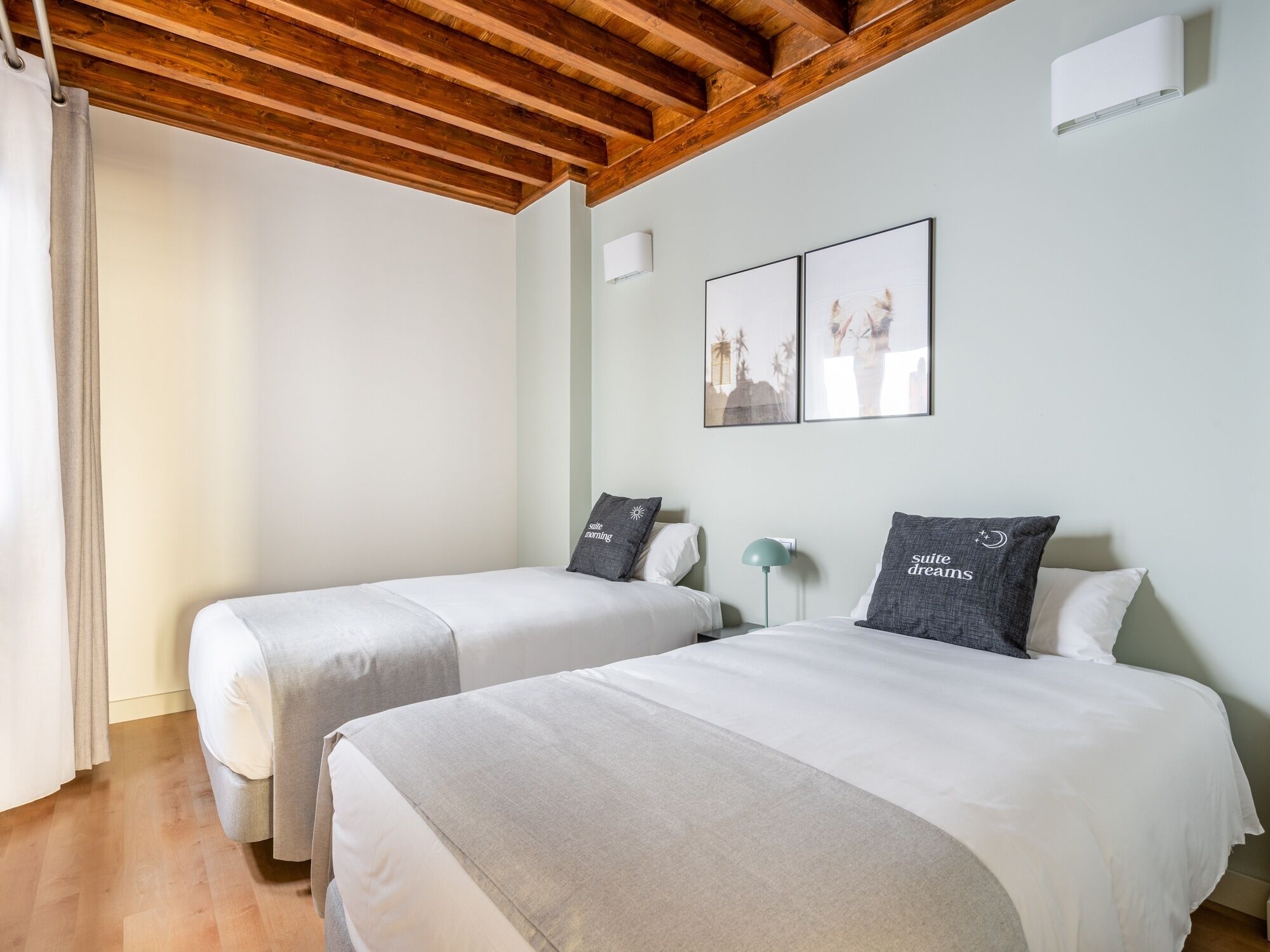 Un dormitorio con dos camas, techo de madera, una mesita de noche con lámpara y dos cuadros enmarcados en la pared.