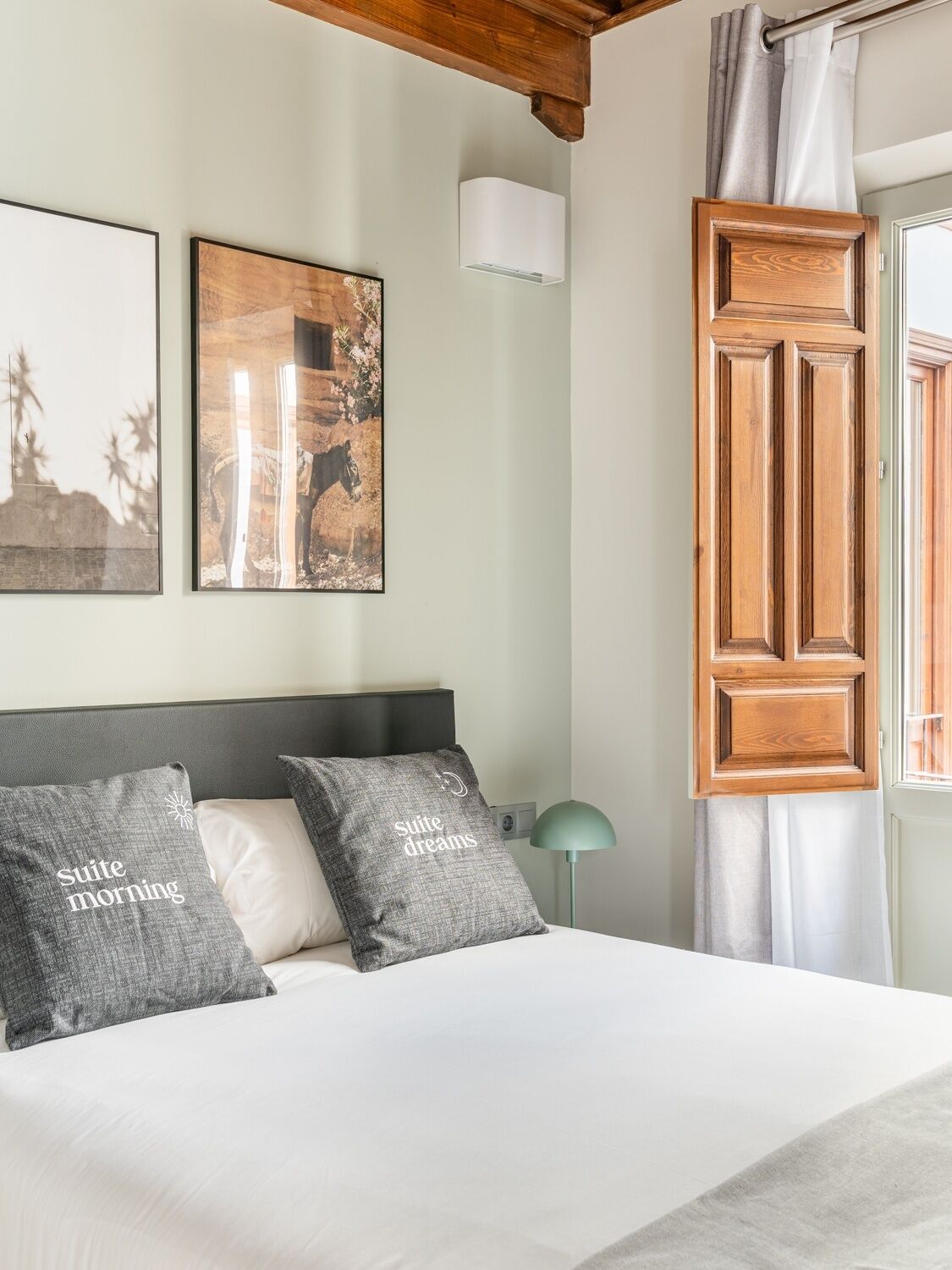 Un dormitorio con cama, dos almohadas, una mesita de noche, lámpara, ventana con cortinas y dos obras de arte colgadas en la pared.