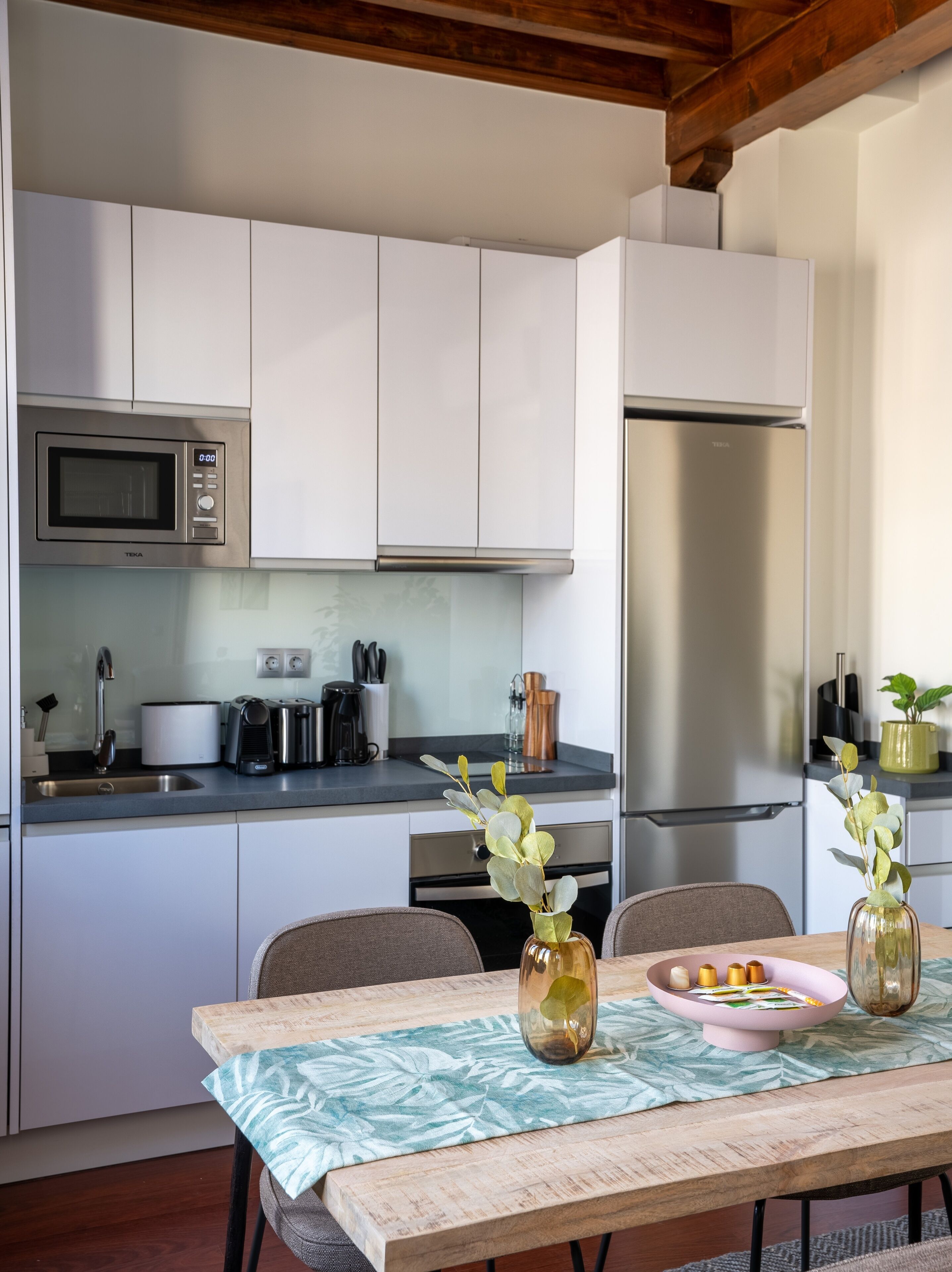 Una cocina moderna con armarios blancos, electrodomésticos de acero inoxidable y un comedor con una mesa dispuesta para dos personas.