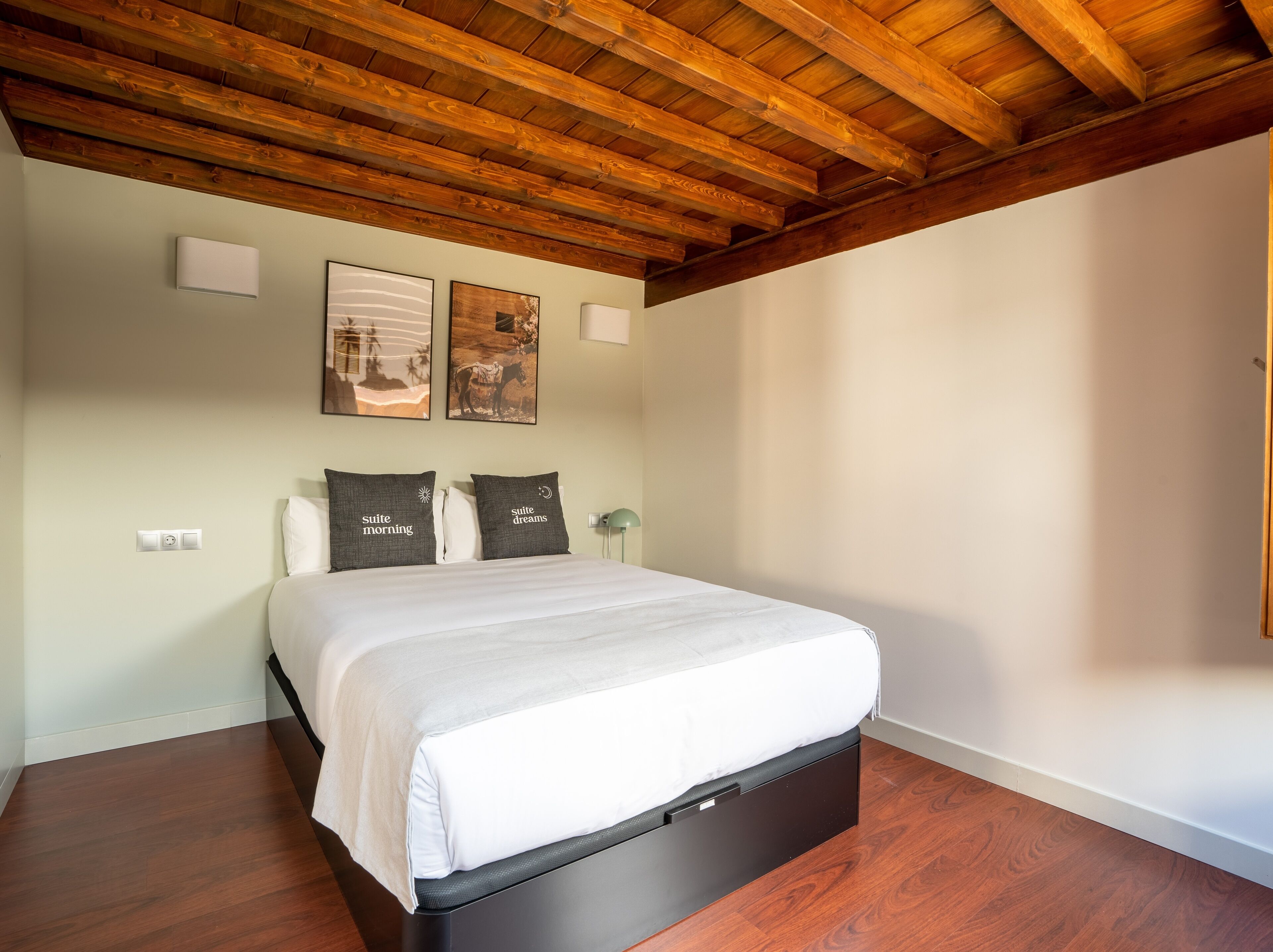 A bedroom with a wooden ceiling, a bed with a white mattress and dark headboard, two pillows with text, and two wall-mounted lights.