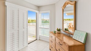 Une pièce dotée d’une porte-fenêtre coulissante donnant sur un balcon, d’une commode en bois avec des tiroirs, d’un miroir et d’un cadre décoratif posé sur la commode.