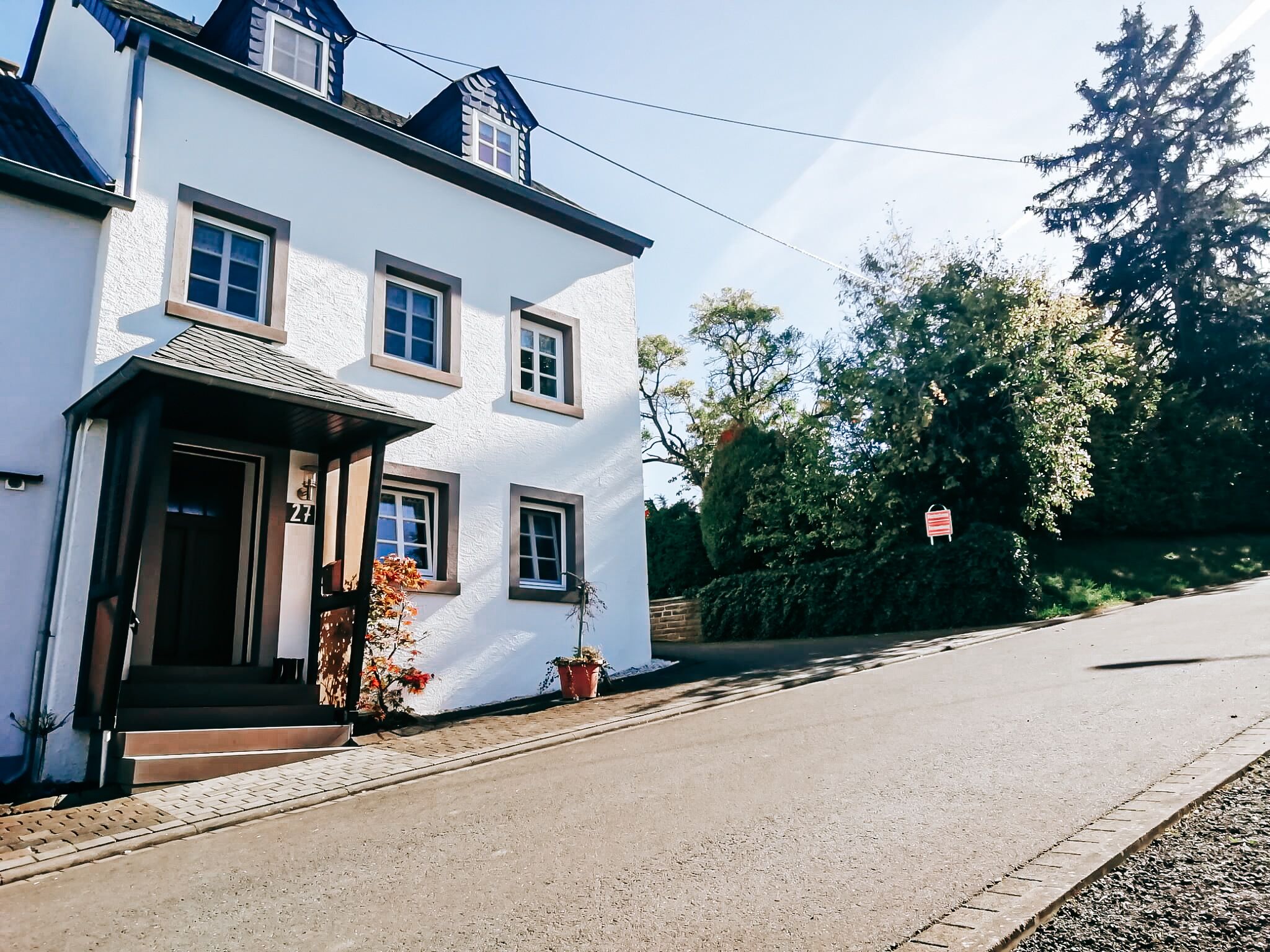 Ein weißes Gebäude mit der Hausnummer 27, einer Veranda mit Stufen und einer Straßenszene mit Bäumen und einer Straße.