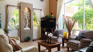 A living room with a sofa, armchairs, a coffee table, and a TV. There are plants, a decorative wall piece, and a window with curtains. - Low Season rate!
 Cozy 3 house, close to the beach, w/private pool and BBQ (Playa del Carmen)