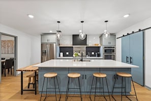 Une cuisine moderne dotée d’un grand îlot central, de tabourets de bar, d’appareils électroménagers en acier inoxydable et de luminaires suspendus.